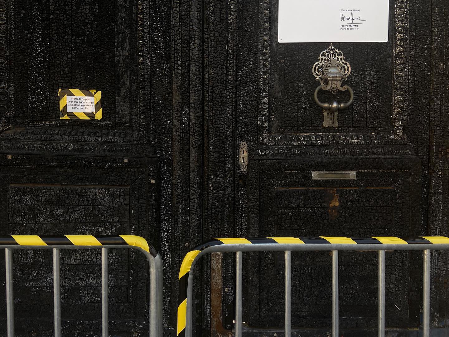 charred and blackened doors of bordeaux city hall. letter taped to the door with black and yellow hazard tape on railings in front