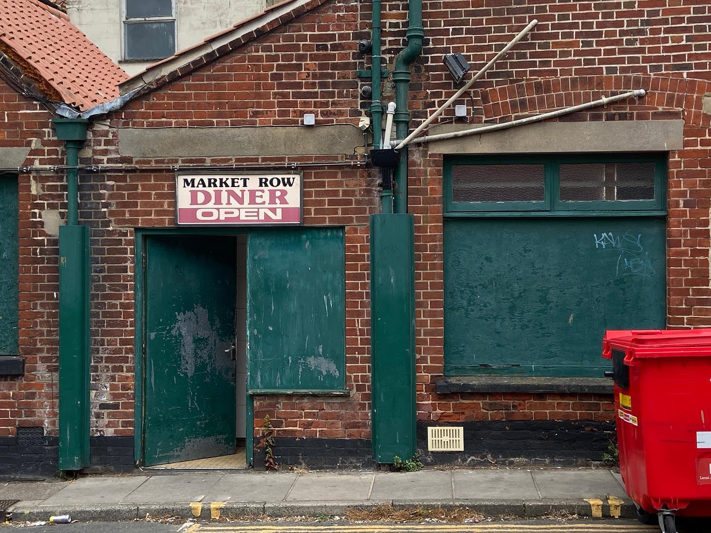 brown brick building with green shutters and doors. to the right is a red biffa bin, and above the door (which is ajar) a sign with red paint saying MARKET ROW DINER OPEN