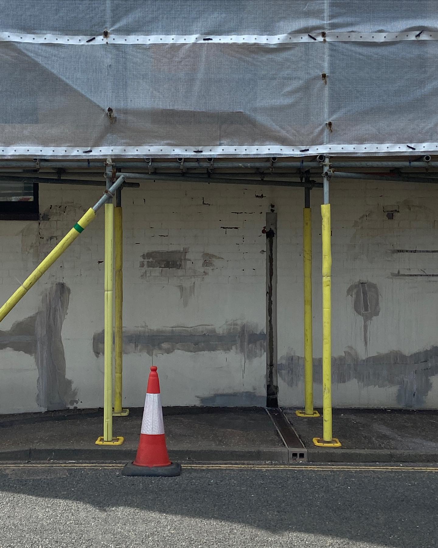 yellow foam scaffold with white netting, over cream and grey wall. a solitary traffic cone