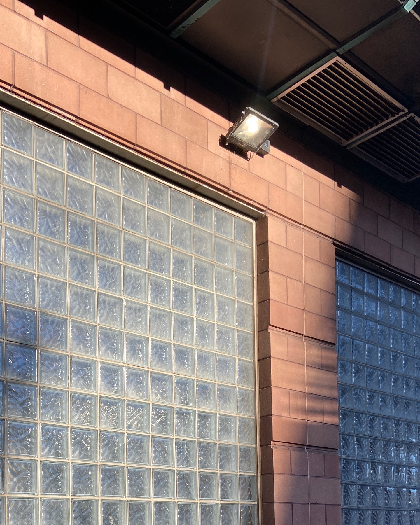 light pink brick with glass brick window. sunlight and soft shadows glinting off floodlight above. grate in ceiling